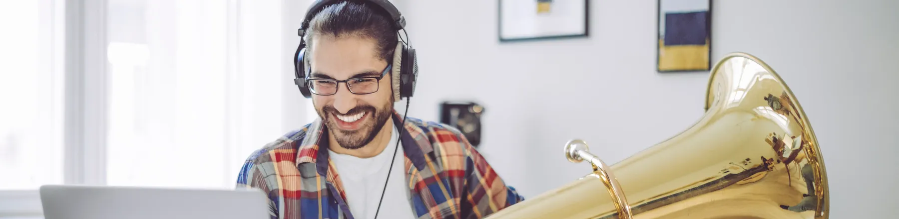 Un uomo sorridente con le cuffie è seduto a una scrivania davanti al suo laptop con una grande tuba tra le braccia, durante una lezione di musica online.