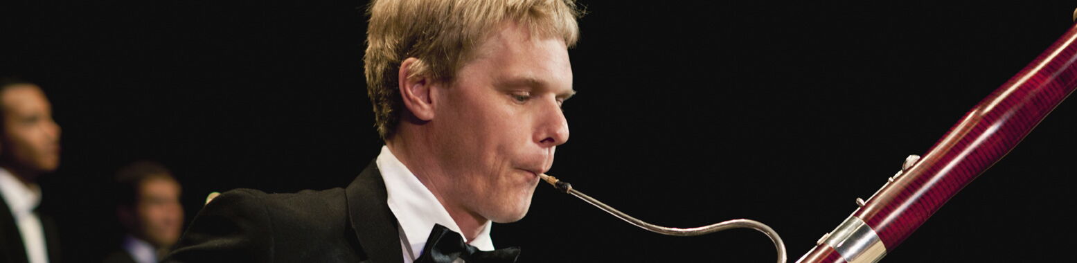 Ein blonder Musiker in Konzertkleidung spielt im Profil ein Fagott vor schwarzem Hintergrund.