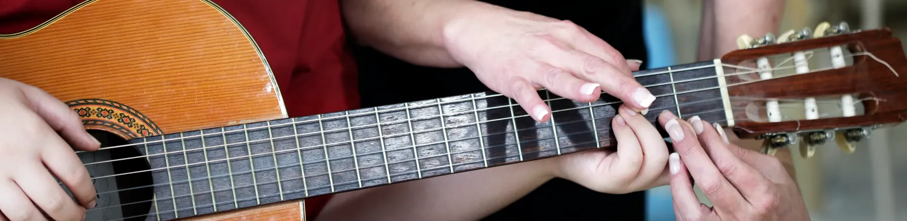 Nahaufnahme von Händen auf dem Griffbrett einer Akustikgitarre; eine Erwachsene hilft einem Kind, die Saiten korrekt zu greifen.