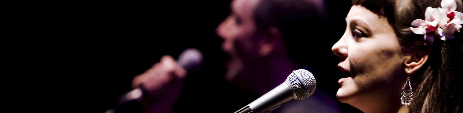 Profilansicht einer Sängerin mit Blumenschmuck im Haar am Mikrofon; im Hintergrund singt unscharf ein männlicher Partner mit.