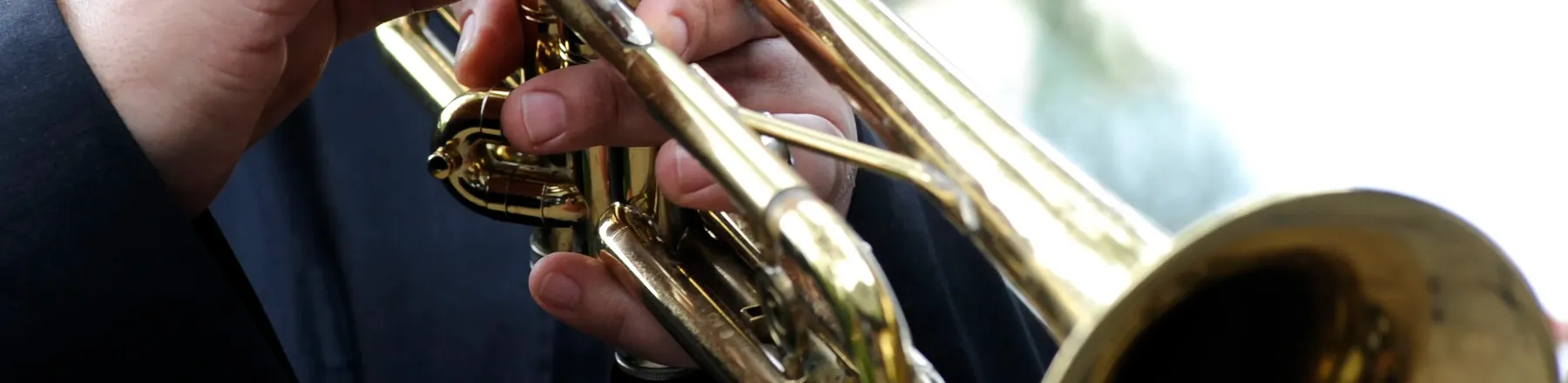 Nahaufnahme der Hände eines Musikers im dunklen Anzug, der eine goldene Trompete spielt; die Finger liegen auf den Ventilen.
