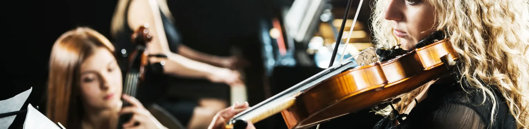 Eine Geigerin mit blonden Locken spielt konzentriert im Scheinwerferlicht; im unscharfen Hintergrund begleiten sie eine Cellistin und eine Pianistin.