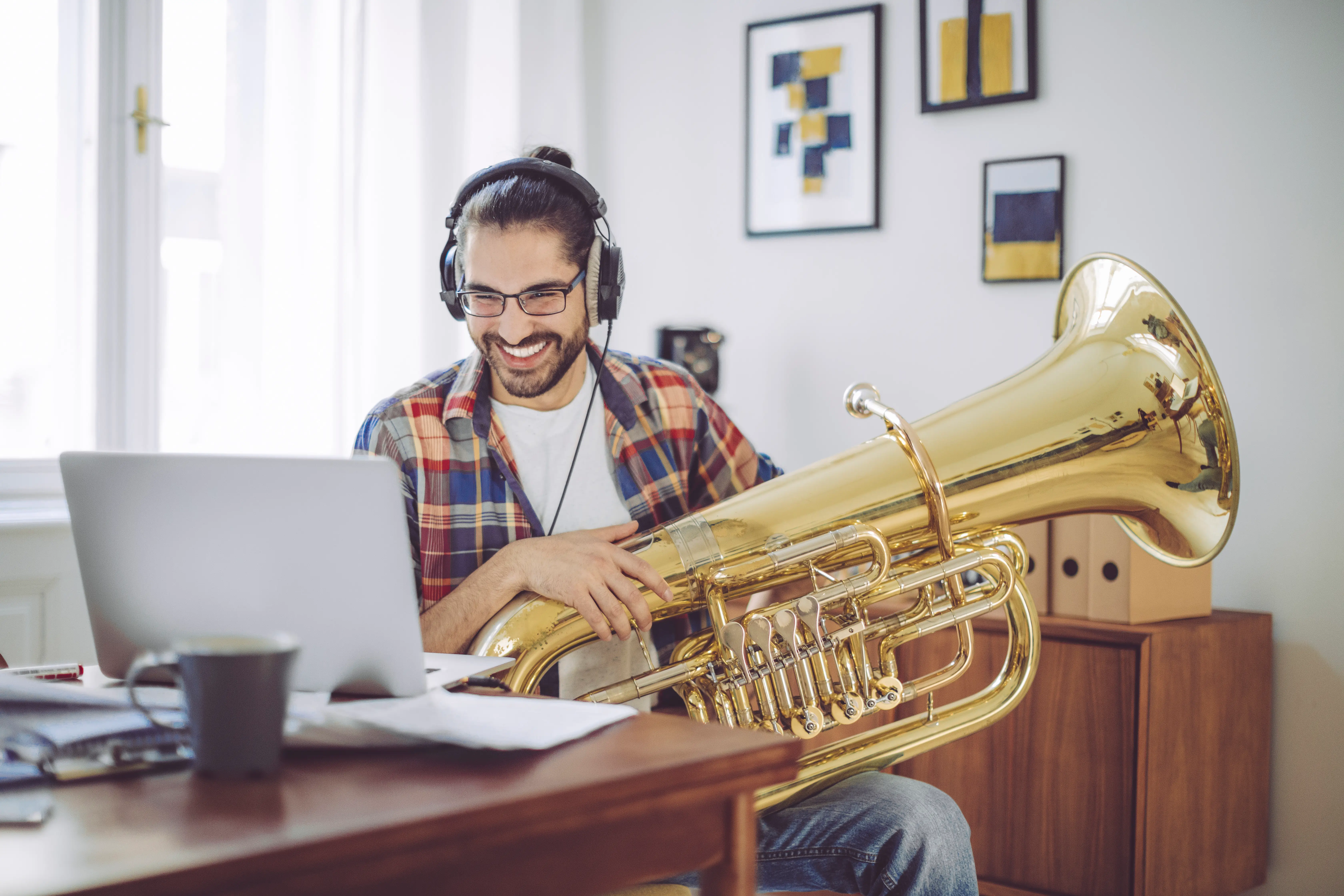 Un uomo sorridente con le cuffie è seduto a una scrivania davanti al suo laptop con una grande tuba tra le braccia, durante una lezione di musica online.