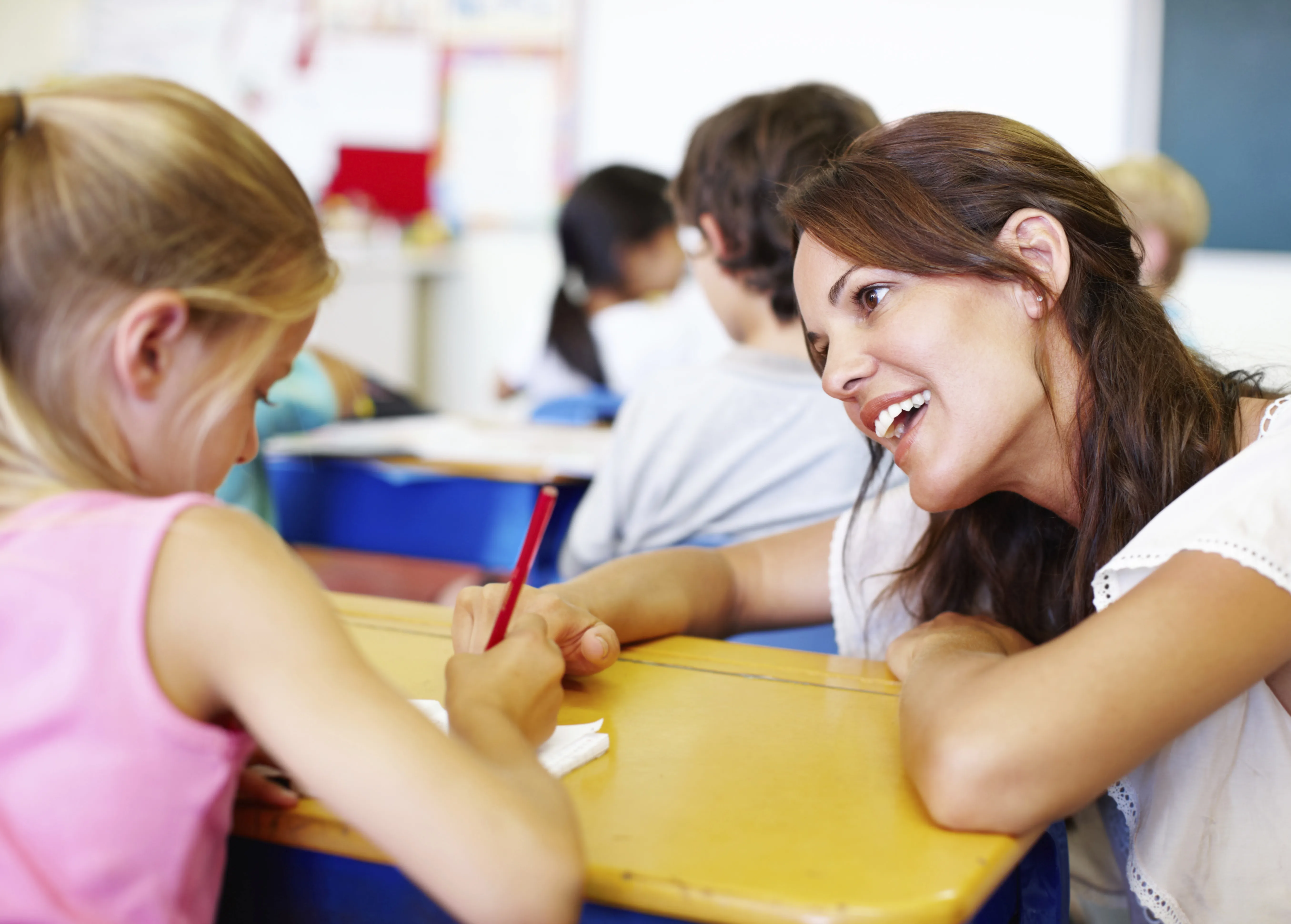 Eine freundlich lächelnde Lehrerin beugt sich im Klassenzimmer zu einer Schülerin am Pult hinunter.