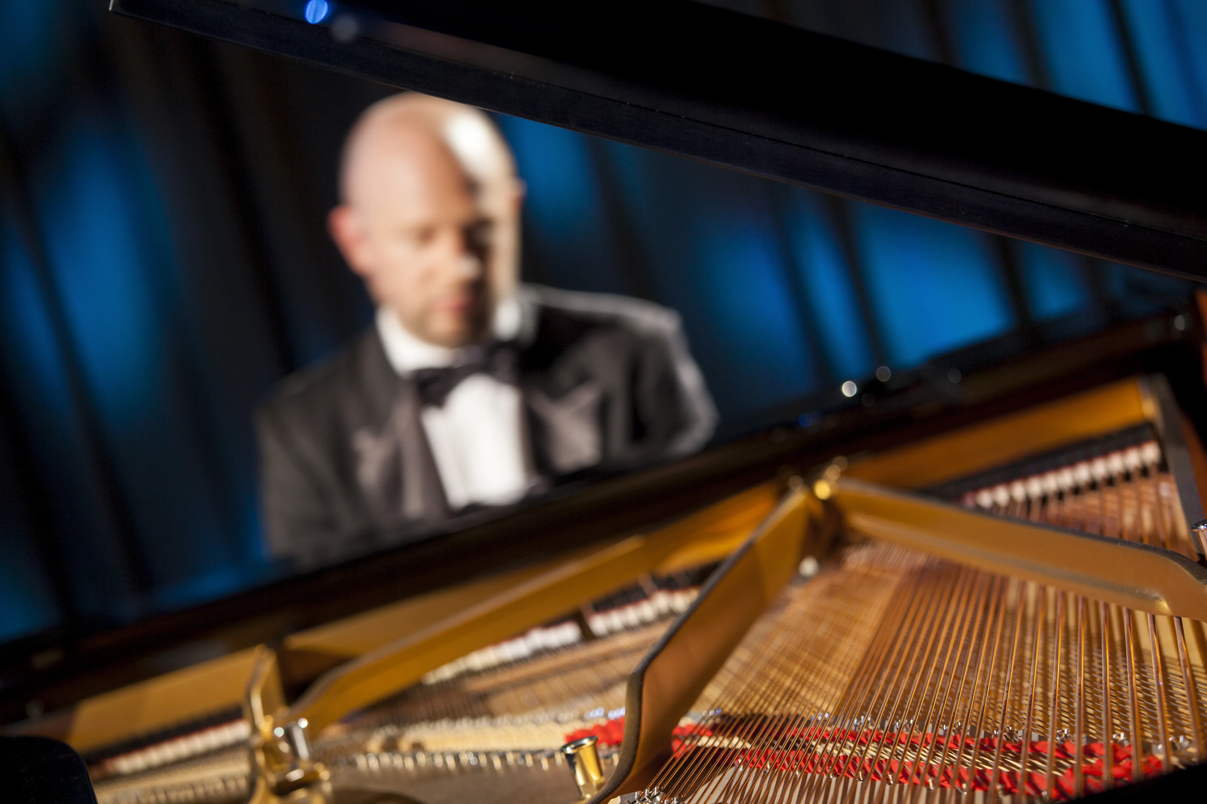 Blick in das Innere eines Klaviers mit Fokus auf die goldenen Saiten und den Rahmen; im Hintergrund ist verschwommen ein Pianist im Smoking zu erkennen.