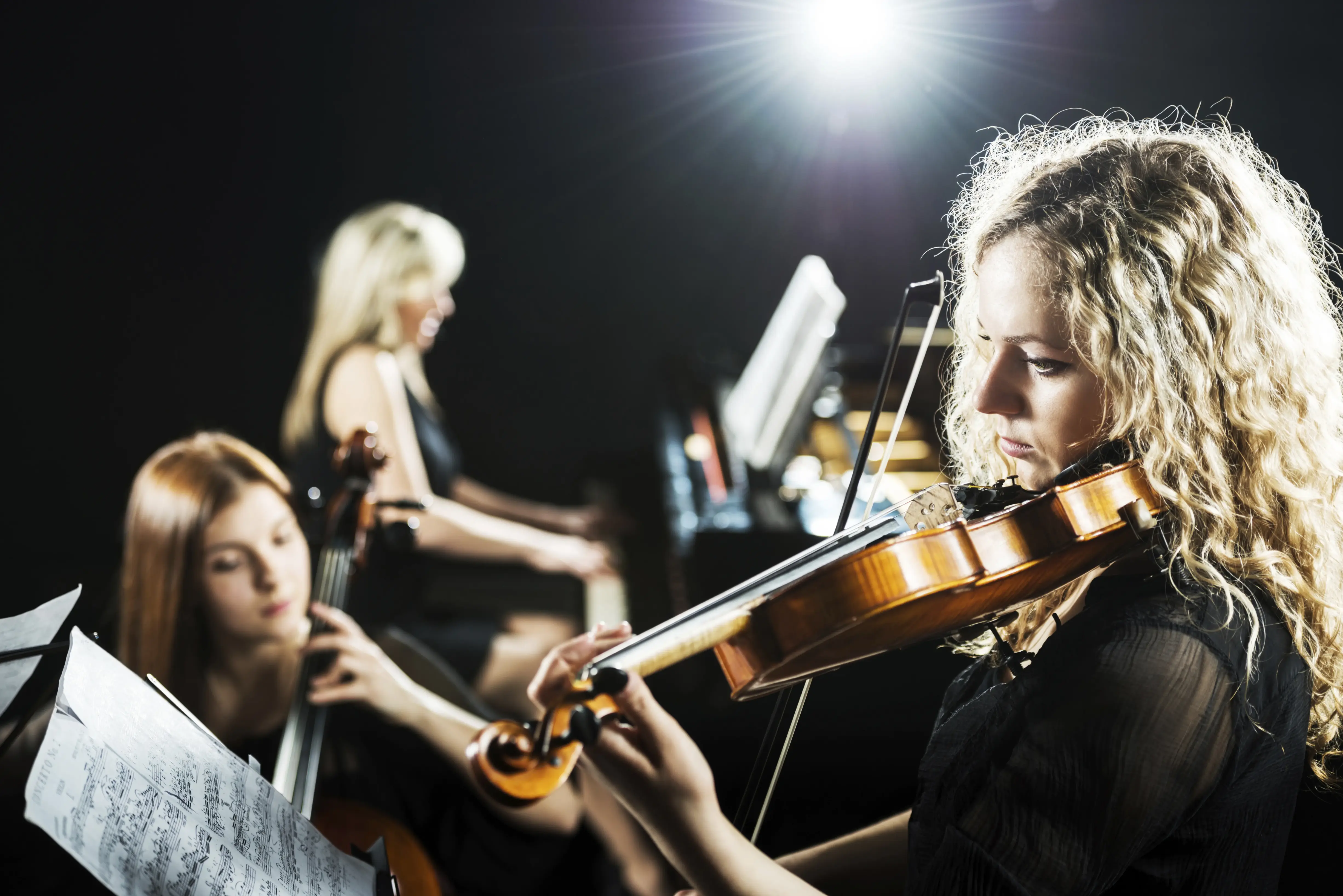 Eine Geigerin mit blonden Locken spielt konzentriert im Scheinwerferlicht; im unscharfen Hintergrund begleiten sie eine Cellistin und eine Pianistin.